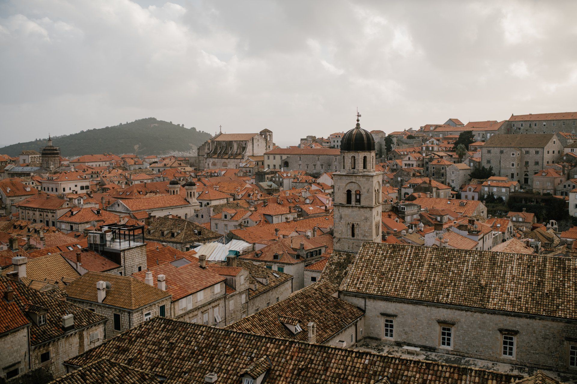 Vista de numerosos tejados de teja rojiza y edificios de piedra, con una torre de cúpula oscura, una colina al fondo y cielo nublado.